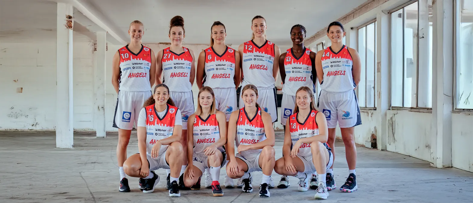Eine Gruppe von acht Frauen in orangefarbenen und weißen Basketballtrikots mit der Aufschrift 'ANGELS' posiert in einer Lagerhalle.
