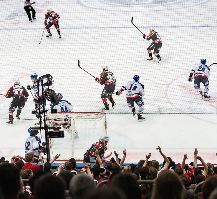 Eine dynamische Eishockeyszene mit Spielern in Aktion auf dem Eis, während die jubelnde Menge im Hintergrund zu sehen ist.