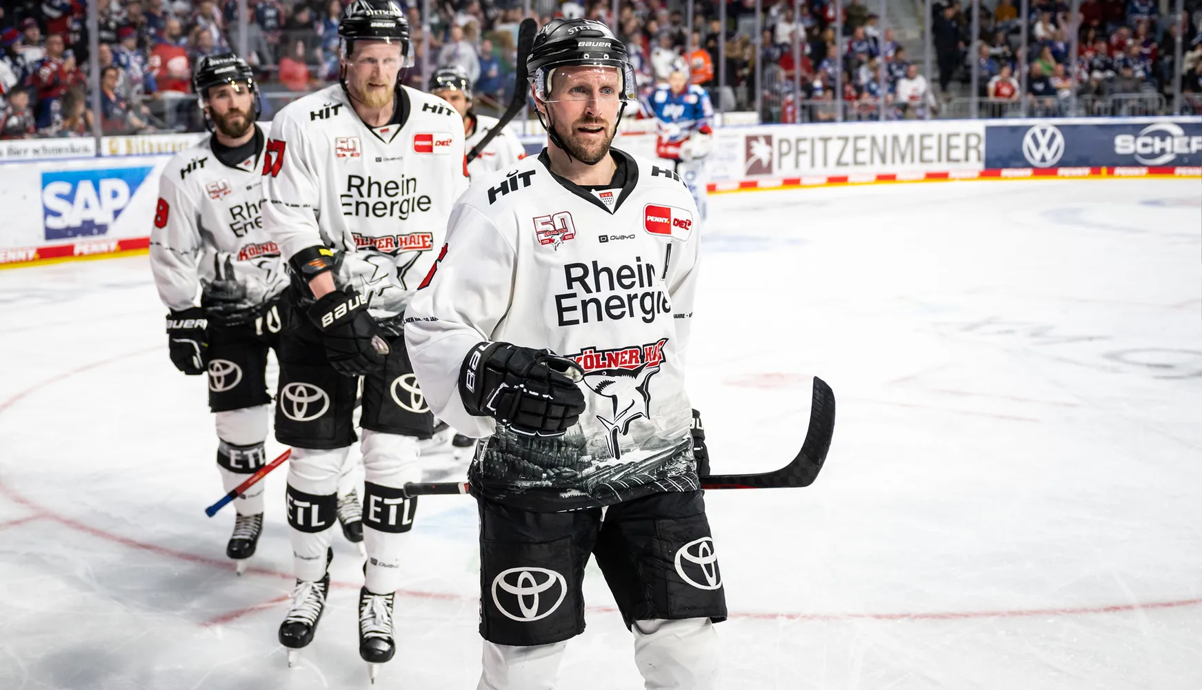 Drei Eishockeyspieler in weißen Trikots mit verschiedenen Nummern stehen auf dem Eis während eines Spiels.
