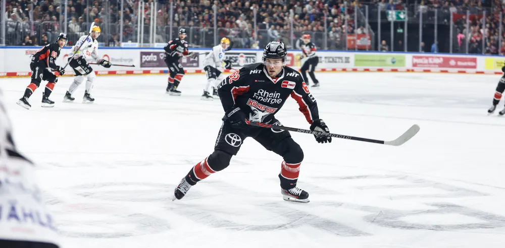 Ein Eishockeyspieler in einem schwarzen Trikot mit roten und weißen Akzenten, der mit einem Schläger auf dem Eis spielt, während die Zuschauer im Hintergrund jubeln.