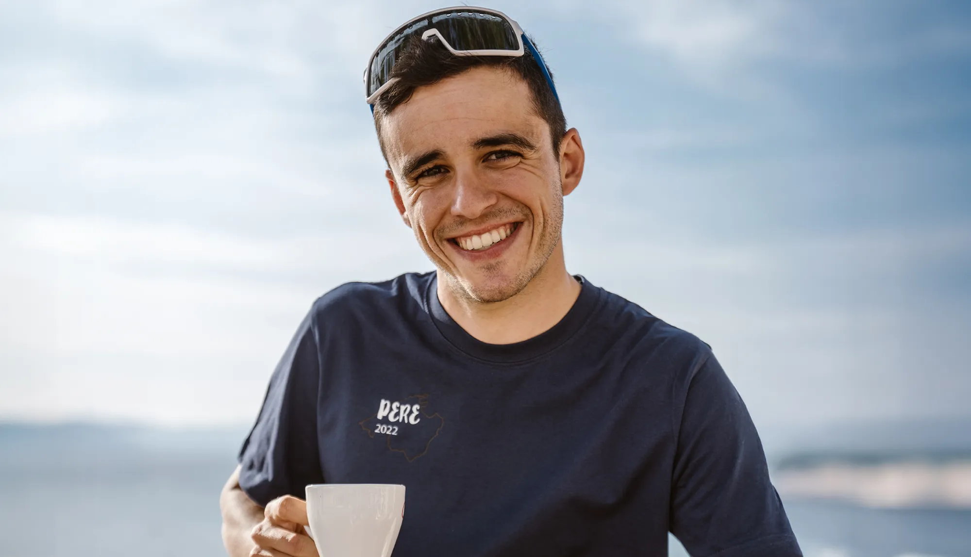 Ein junger Mann lächelt in einem blauen T-Shirt mit dem Aufdruck 'PERE 2022' und hält eine Tasse in der Hand, während er vor einem klaren Himmel steht.