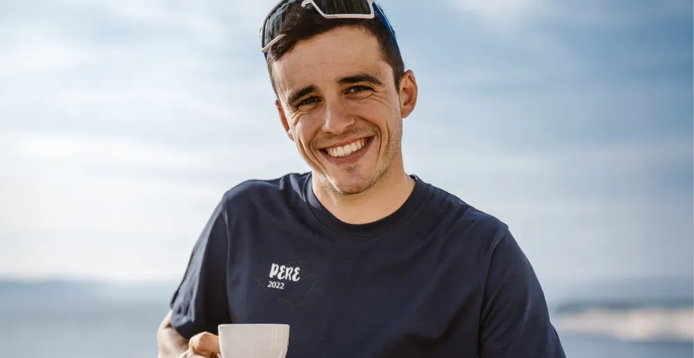 Ein sportlicher Mann in einem dunkelblauen T-Shirt mit dem Aufdruck 'PERE 2022', der lächelnd eine Tasse hält, vor einer verschwommenen Küstenlandschaft.