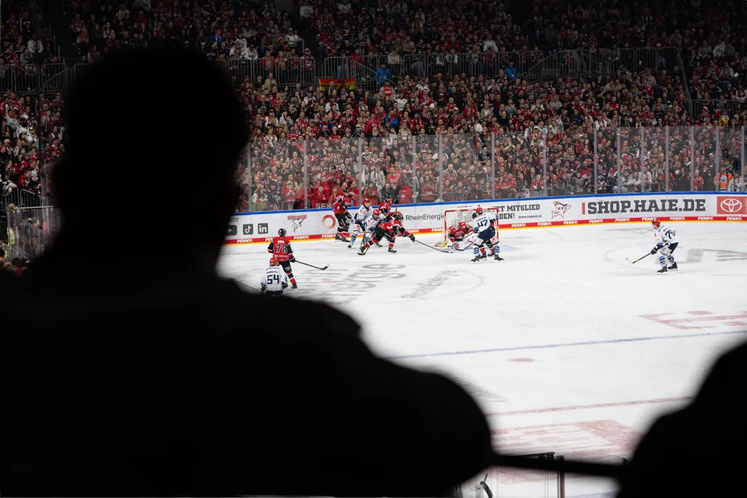 Aus der Zuschauersicht zeigt das Bild ein Eishockeyspiel mit Spielern in roten und blauen Trikots vor einer großen Zuschauermenge.