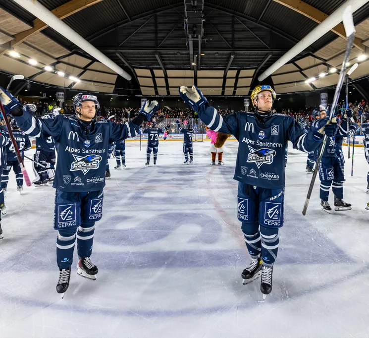 Eishockeyspieler in blauen Trikots mit Sponsorenlogos feiern auf dem Eis, während im Hintergrund das Publikum zu sehen ist.