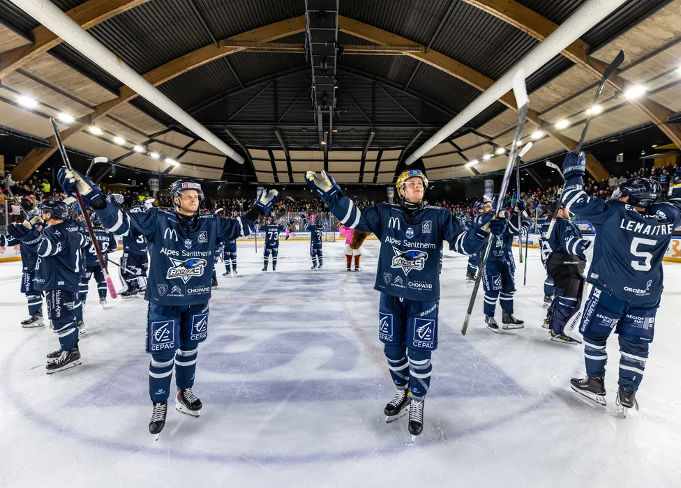 Eishockeyspieler in blauen Trikots stehen auf dem Eis und feiern mit erhobenen Schlägern vor einer jubelnden Menge.