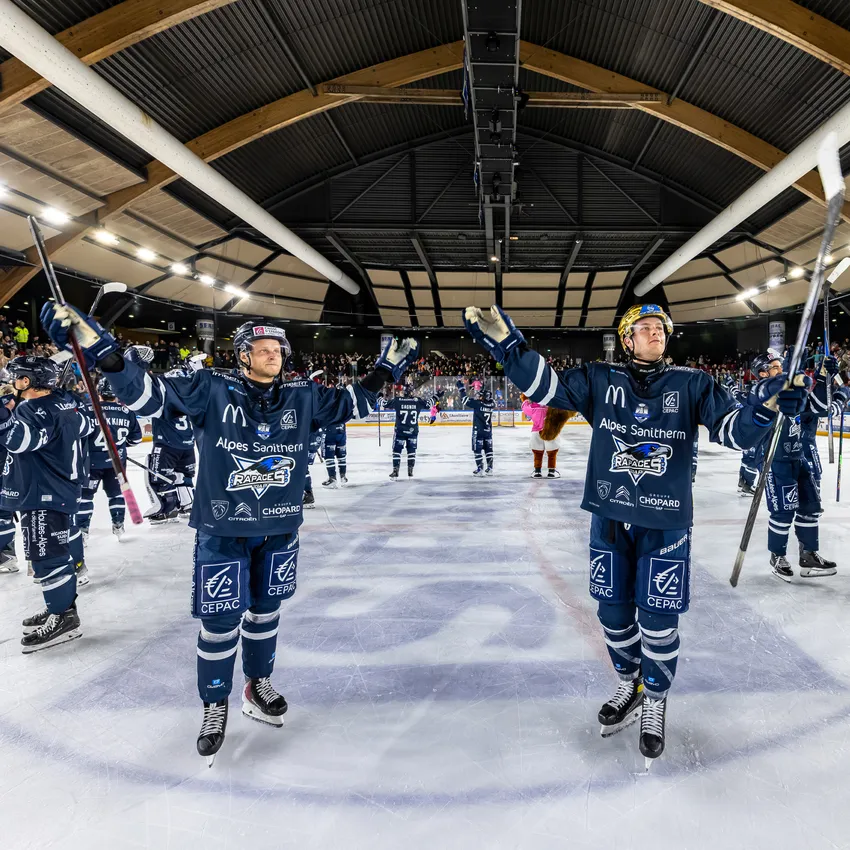 Zwei Eishockeyspieler in blauen Trikots, die auf dem Eis stehen und ihre Schläger heben, während eine jubelnde Menge im Hintergrund zu sehen ist.