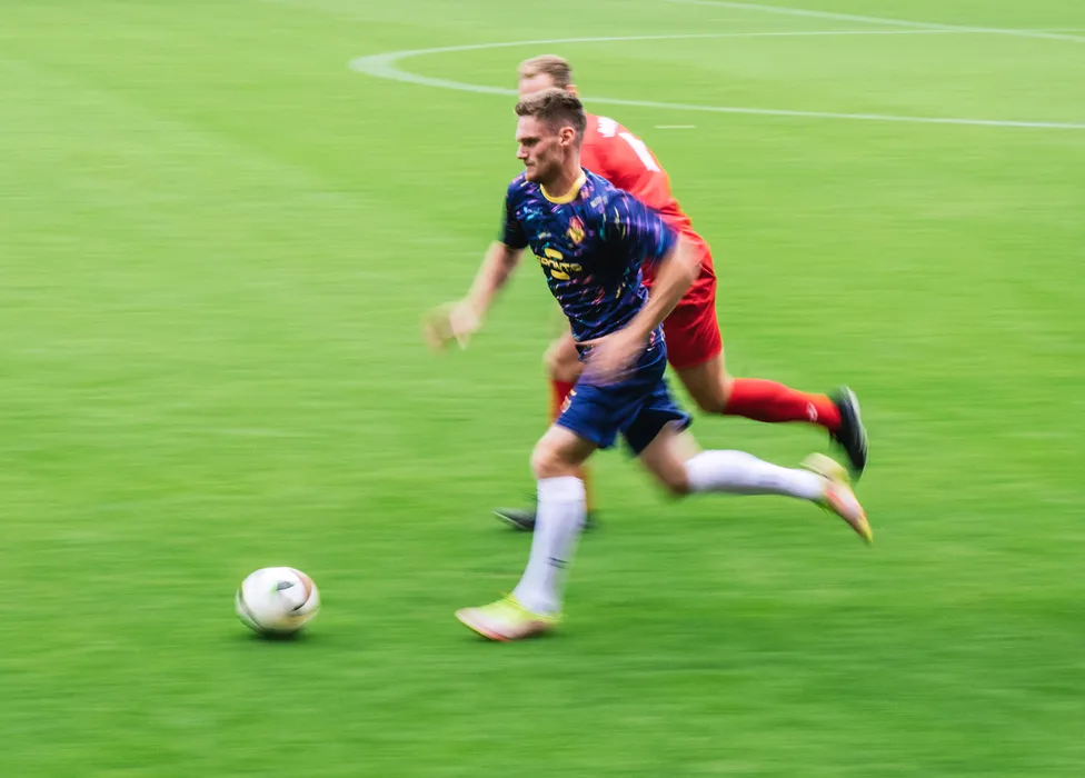 Ein Fußballspieler in einem blauen Trikot mit bunten Mustern läuft schnell über das grüne Spielfeld, während ein anderer Spieler ihm nahe folgt.