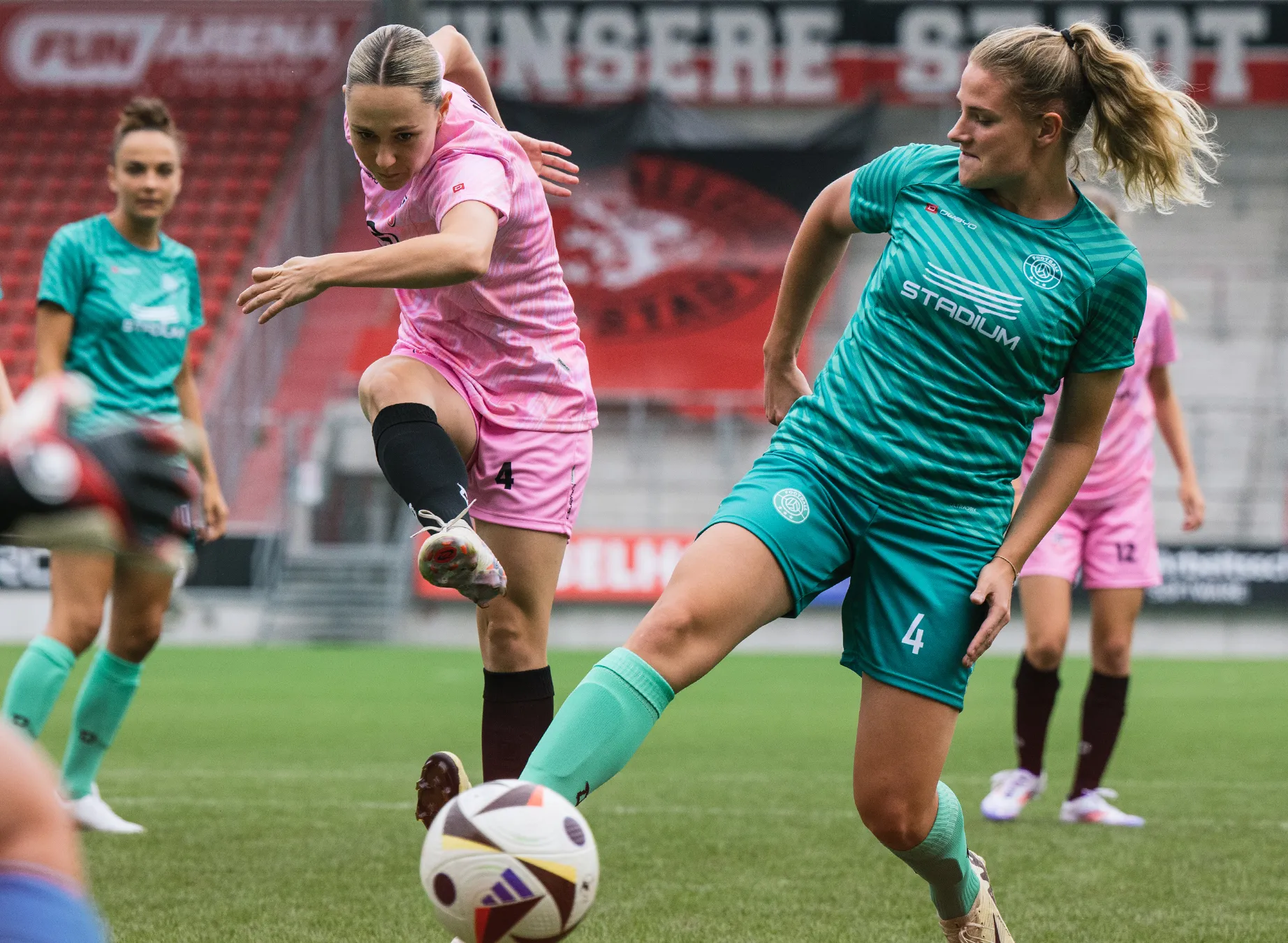 Zwei Fußballspielerinnen in lebhaften Trikots, eine in pink und die andere in grün, spielen auf einem Rasenplatz.