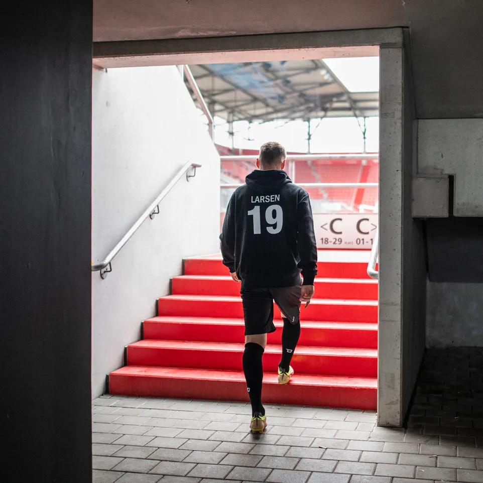 Ein Sportler mit einem schwarzen Kapuzenpullover und Shorts, der auf eine rote Treppe im Stadion zugeht.