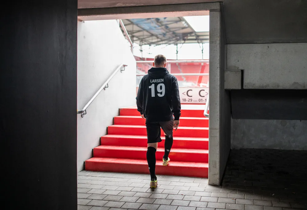 Ein Fußballspieler in einem schwarzen Hoodie mit der Nummer 19 und kurzen Hosen geht die roten Stufen eines Stadions hinauf.