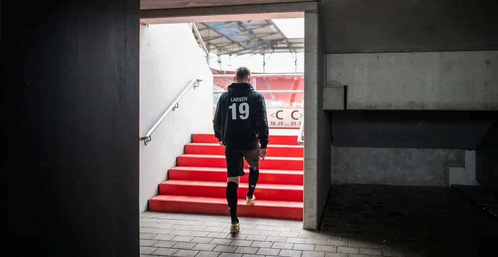 Ein Sportler in einem schwarzen Hoodie mit der Nummer 19 und schwarzen Shorts, der auf eine rote Treppe in einem Stadion zugeht.