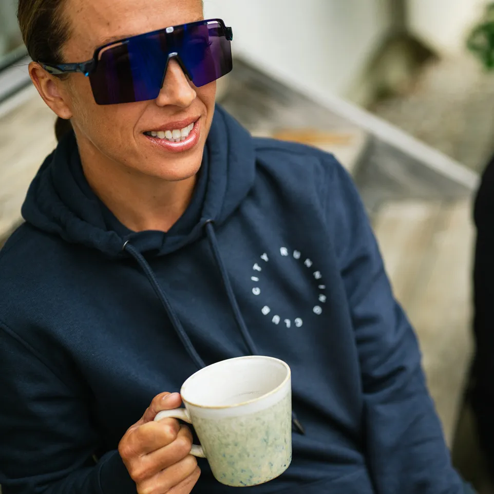 Eine Person trägt einen dunkelblauen Hoodie mit einem Schriftzug und hält eine Tasse in der Hand, während sie in die Kamera lächelt.