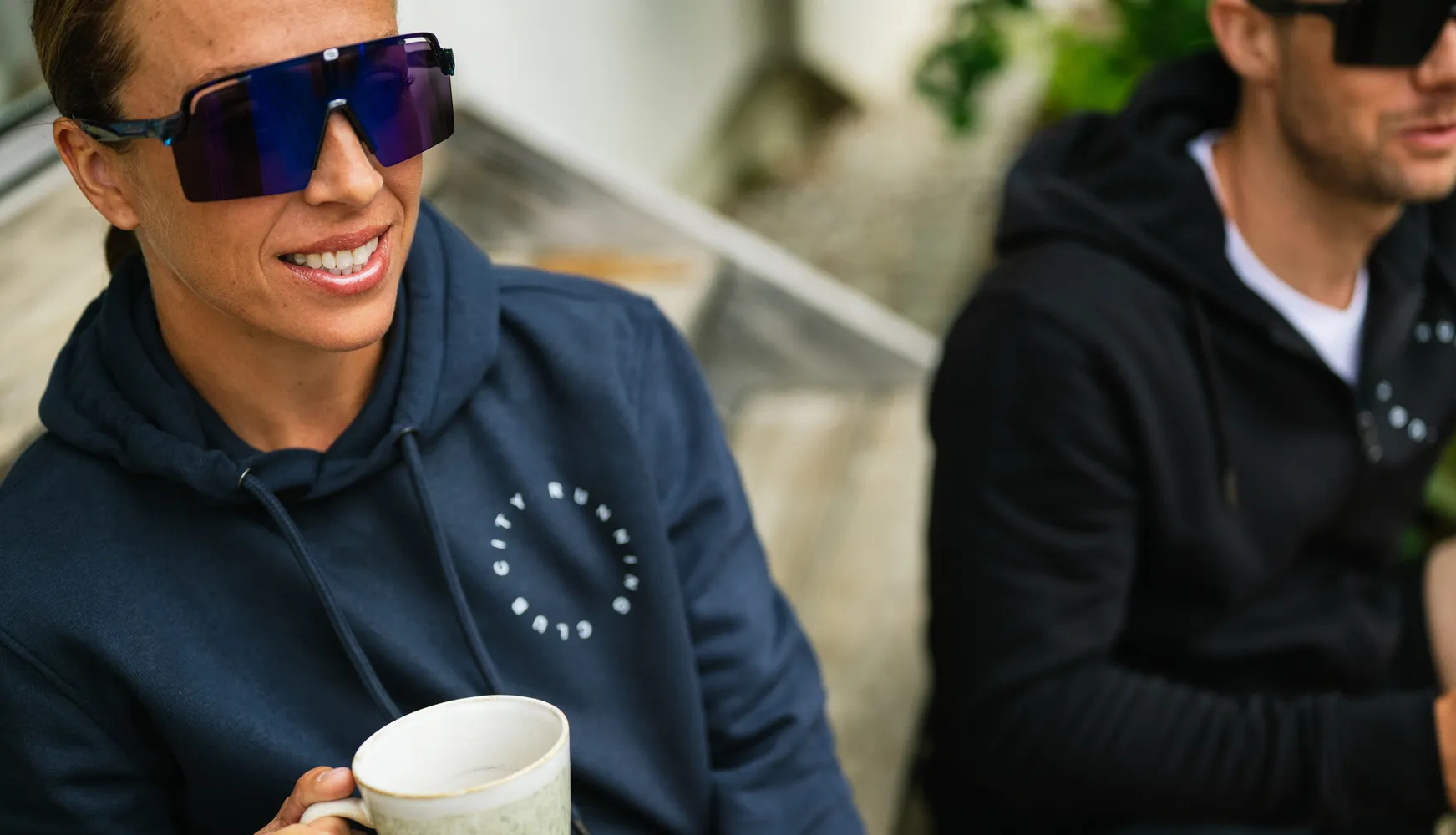 Eine Person in einem blauen Hoodie hält eine Tasse und trägt große Sonnenbrillen, während eine weitere Person im Hintergrund ebenfalls einen Hoodie trägt.