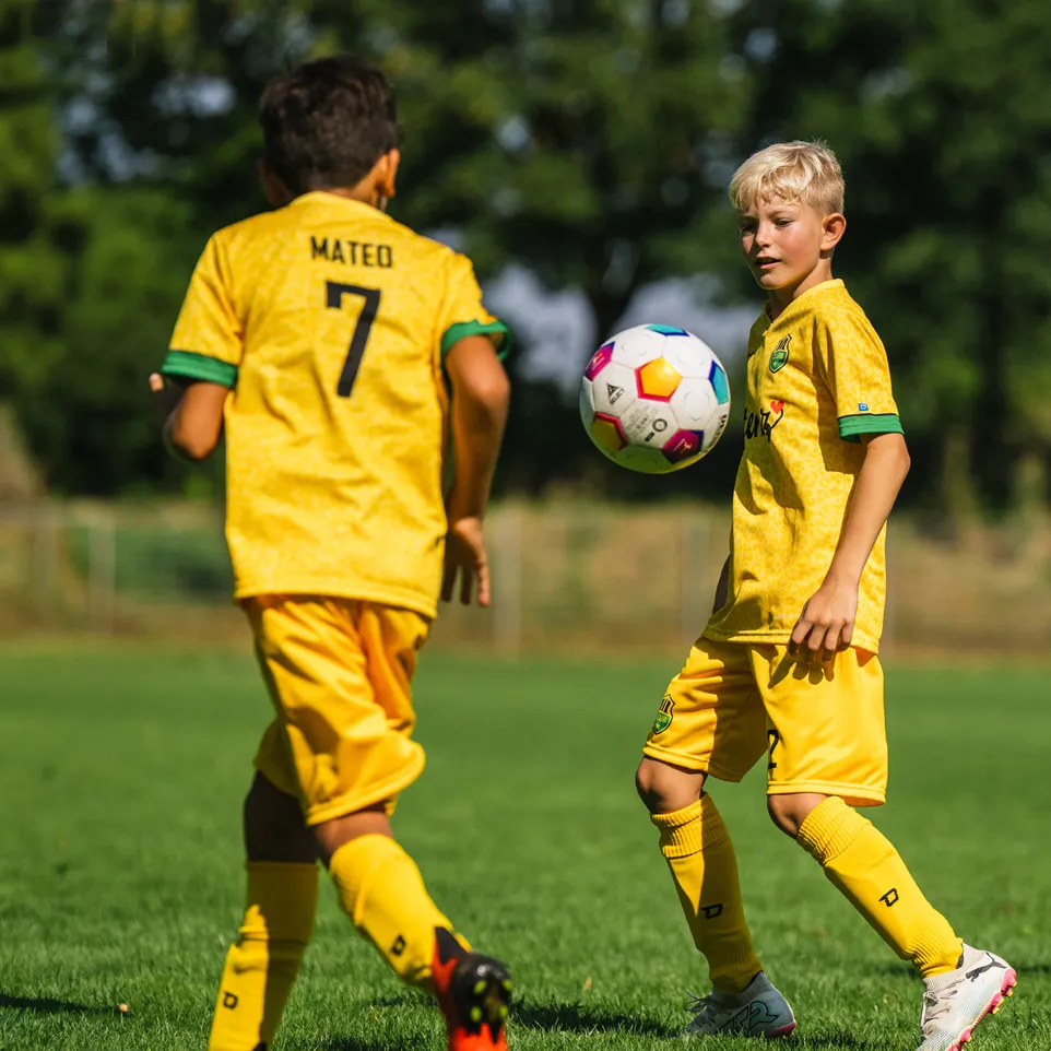 Zwei Kinder in gelben Fußballtrikots und Shorts spielen auf einem grünen Fußballplatz.