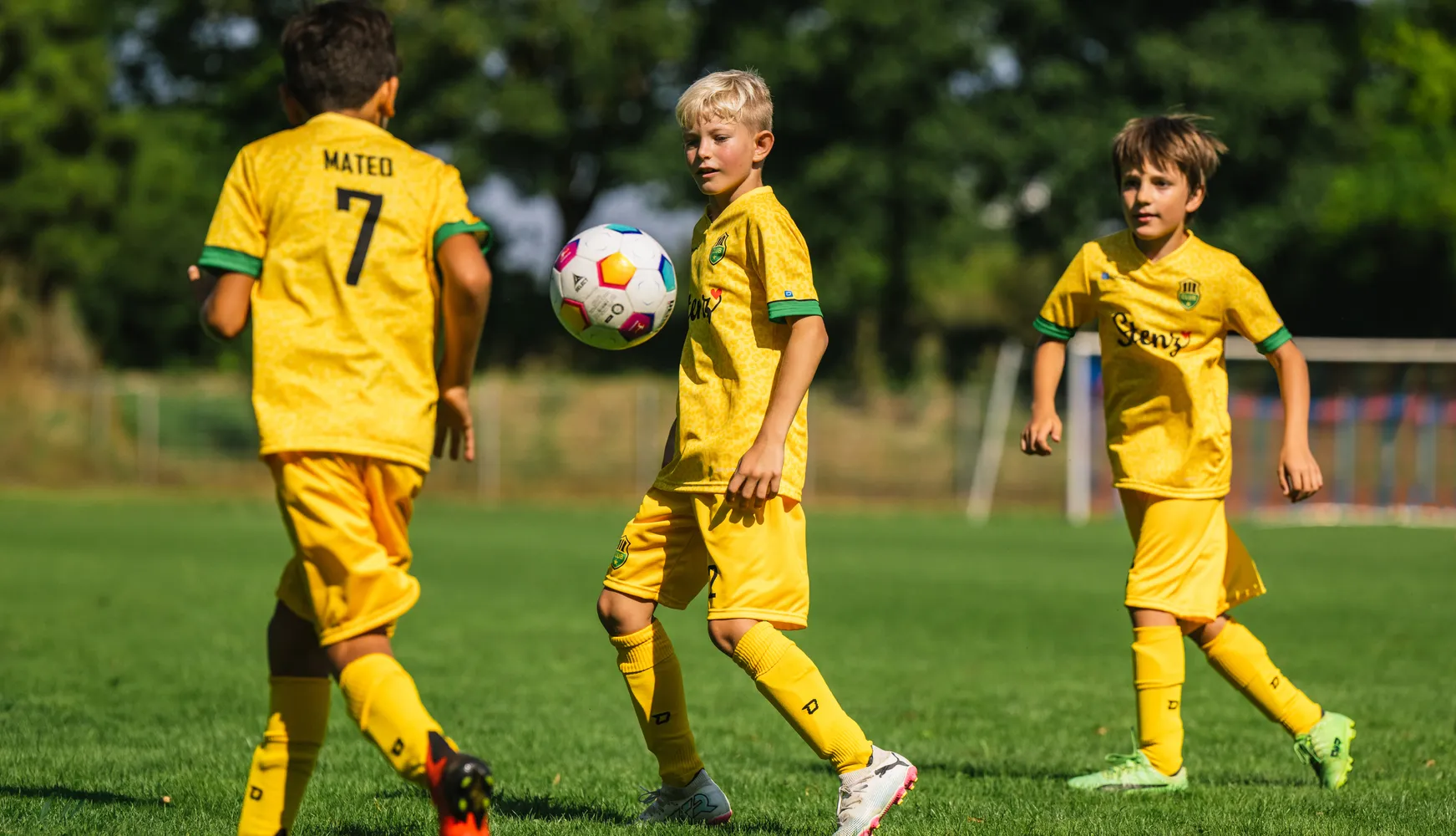Drei Kinder in gelben Fußballtrikots spielen auf einem Rasenplatz mit einem bunten Fußball.