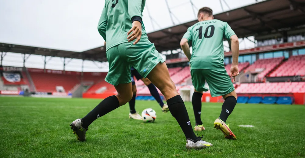 Zwei Fußballspieler in hellgrünen Trikots und schwarzen Socken, die auf einem Rasenfeld in einem Stadion trainieren.