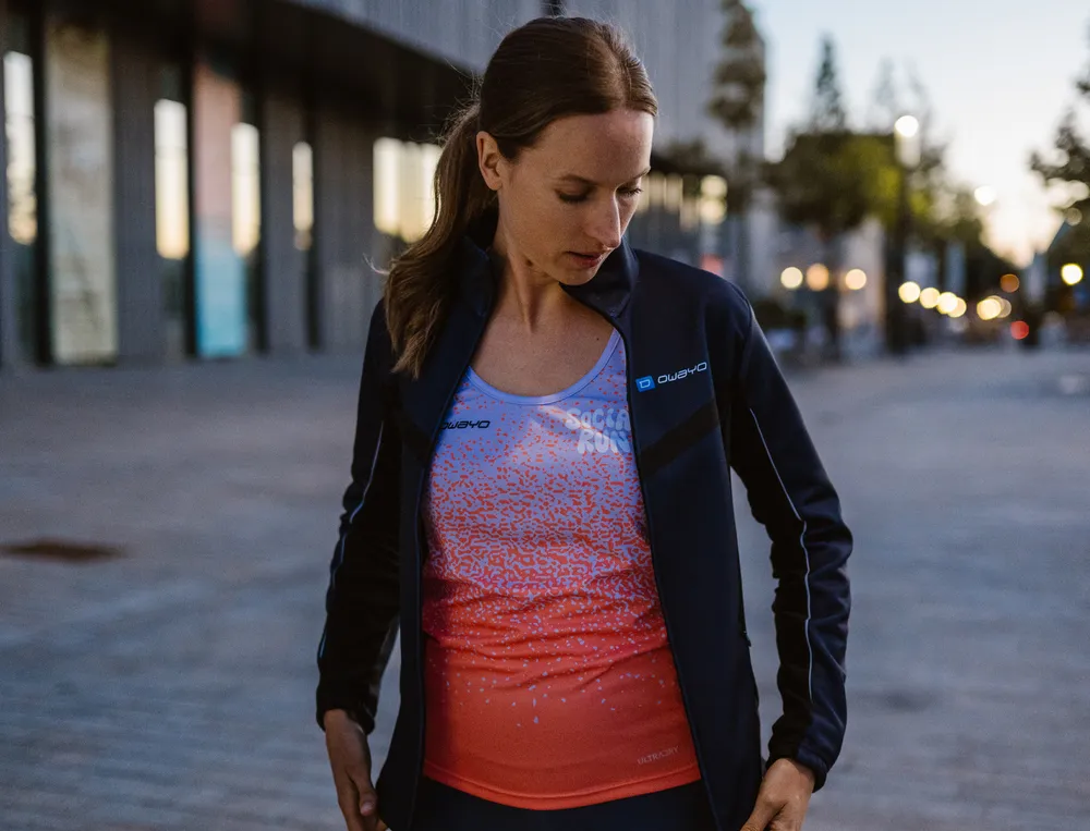 Eine Frau in sportlicher Kleidung, bestehend aus einer leichten Jacke und einem bunten Oberteil, steht in einer urbanen Umgebung und schaut nachdenklich auf ihr Outfit.