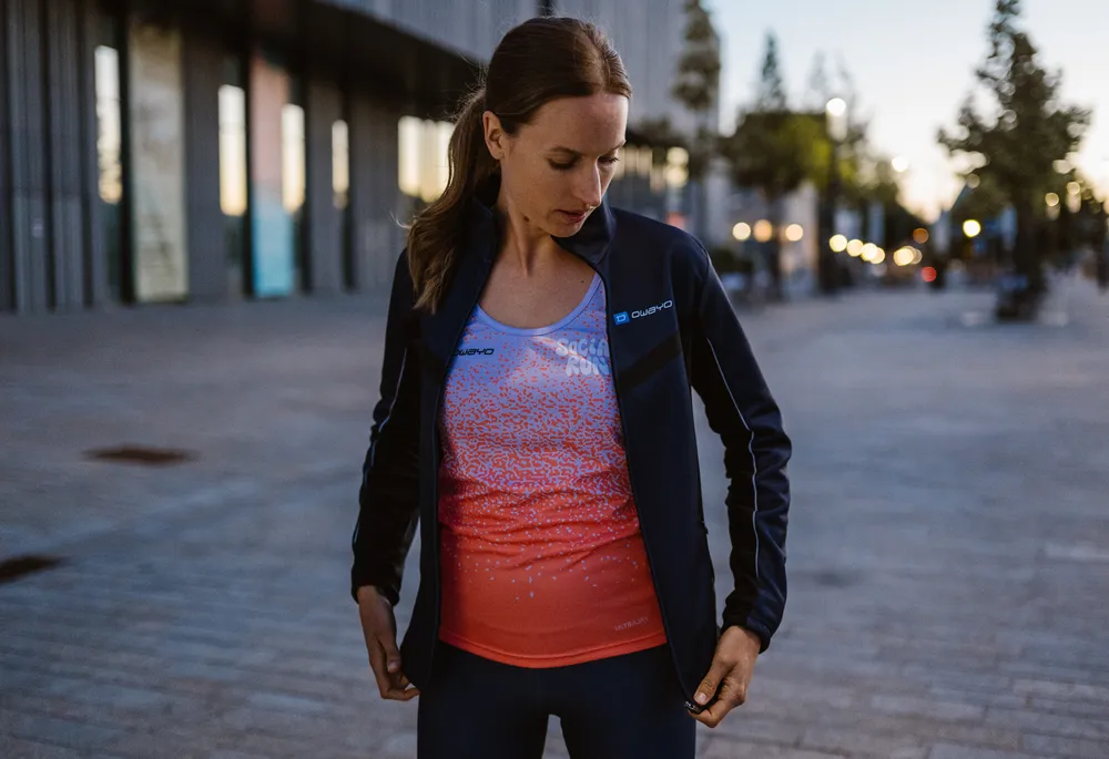 Eine sportliche Frau trägt eine dunkelblaue Jacke über einem farbverlaufenden, ärmellosen Oberteil in Blau und Orange, während sie in einer urbanen Umgebung steht.