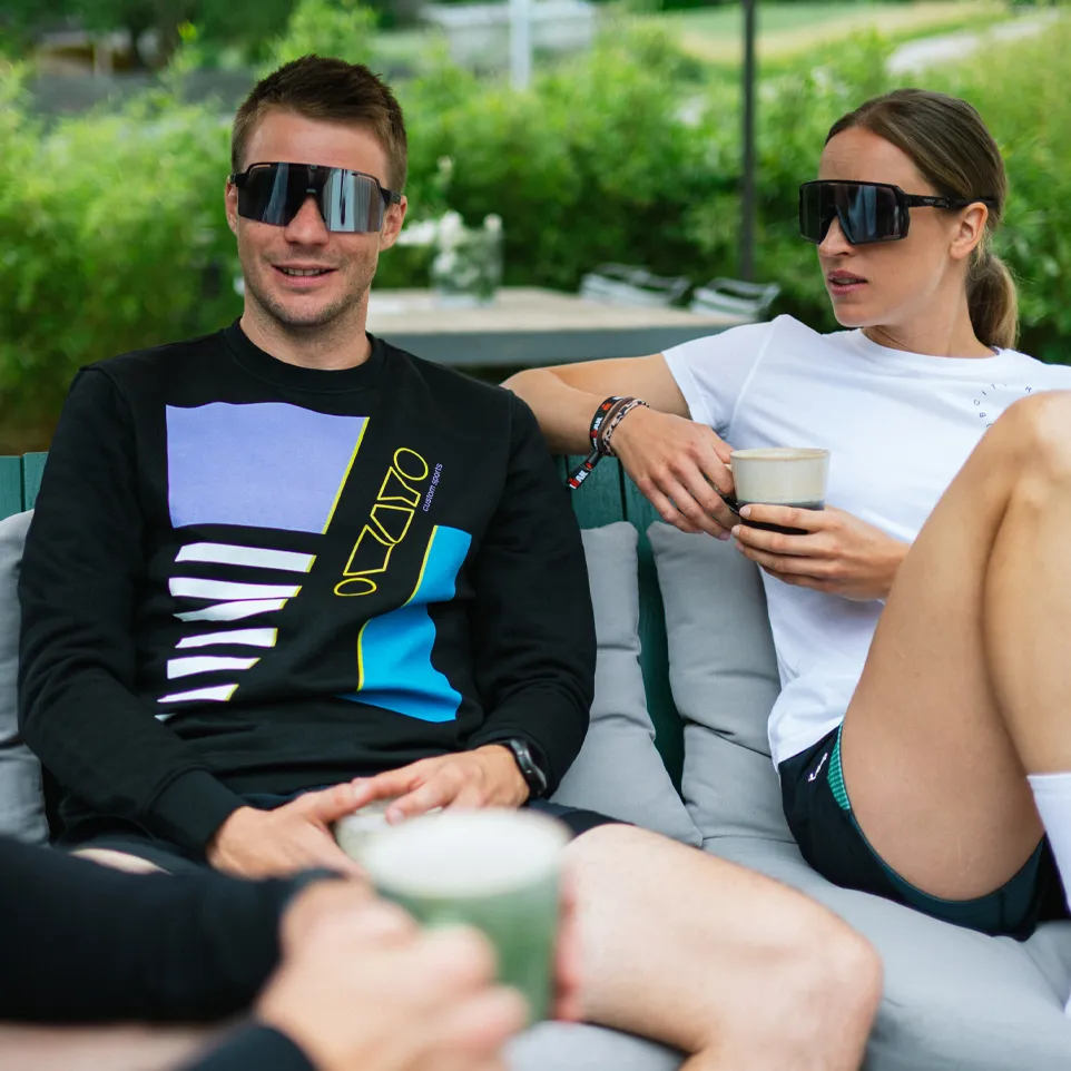 Zwei Personen sitzen auf einer Couch im Freien, der Mann trägt einen schwarzen Pullover mit buntem Design und die Frau ein weißes T-Shirt, beide halten Getränke in den Händen.