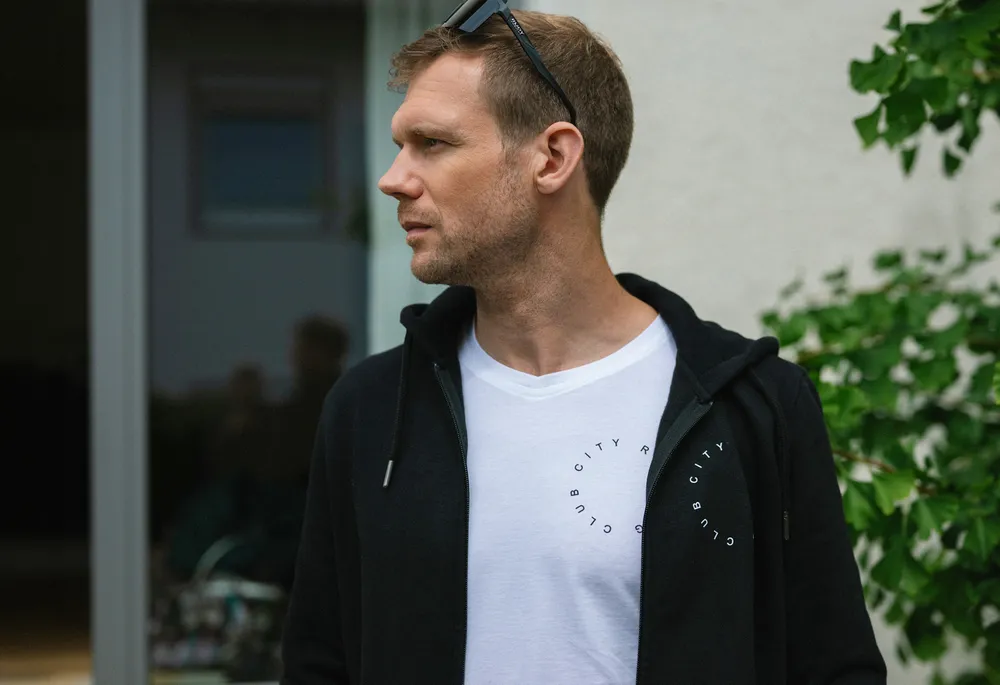 Ein Mann steht seitlich mit einem schwarzen Hoodie und einem weißen T-Shirt mit einem runden Aufdruck.