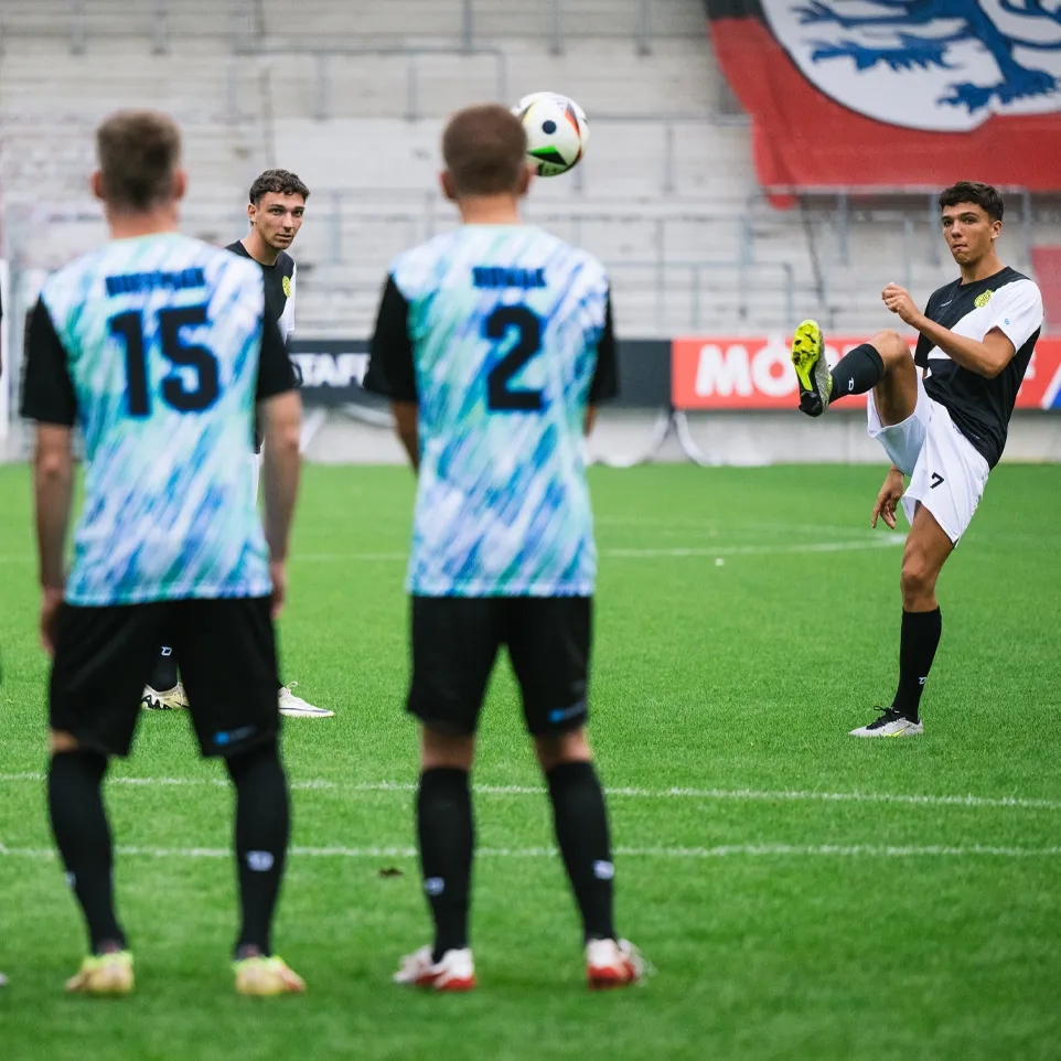 Drei Fußballspieler in bunten Trikots stehen auf dem Platz und schauen auf einen anderen Spieler, der einen Ball jongliert.