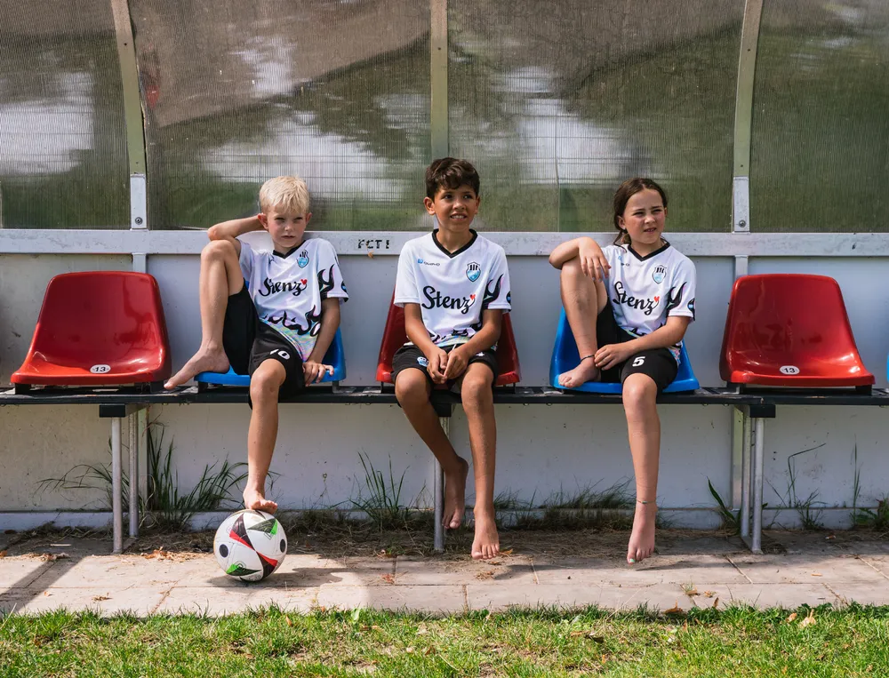 Drei Kinder sitzen auf einer Bank am Spielfeldrand, tragen individuell gestaltete Fußballtrikots und haben einen Fußball vor sich.