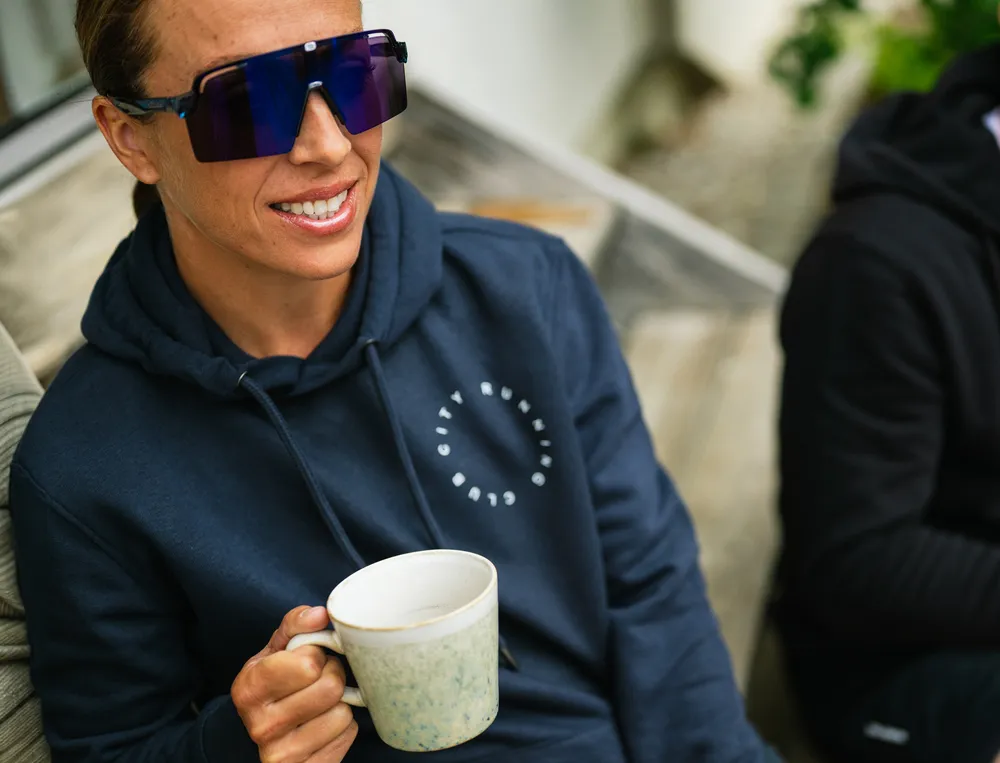 Eine Person in einem navyfarbenen Hoodie mit einem Logo hält eine Tasse Kaffee und trägt große Sonnenbrillen.