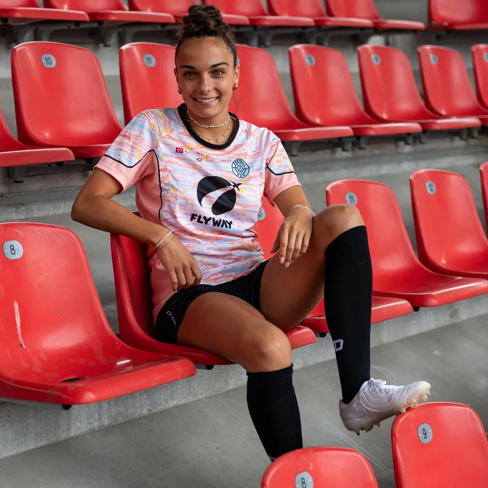 Eine lächelnde Frau sitzt in einem Stadion auf einem roten Sitz, trägt ein farbenfrohes Fußballtrikot und schwarze Socken.