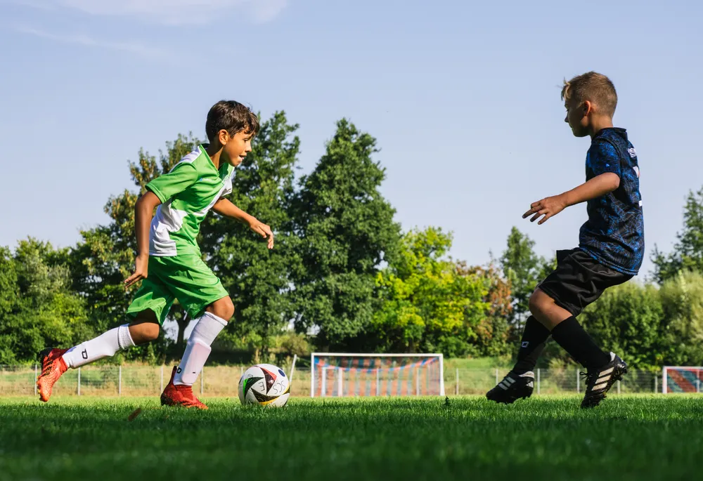 Zwei Kinder spielen Fußball auf einem grünen Platz, einer in einem grünen Trikot mit weißen Akzenten und der andere in einem blauen Shirt, während sie einem Ball nachjagen.