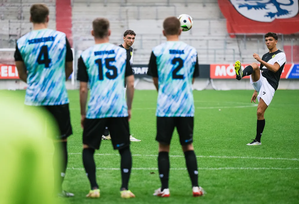 Eine Gruppe von Fußballspielern in bunten Trikots steht auf dem Platz, während ein Spieler einen Freistoß vorbereitet.