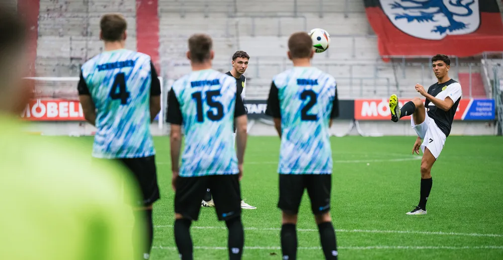 Ein Fußballspieler führt einen Schuss aus, während andere Spieler in bunten Trikots zuschauen.