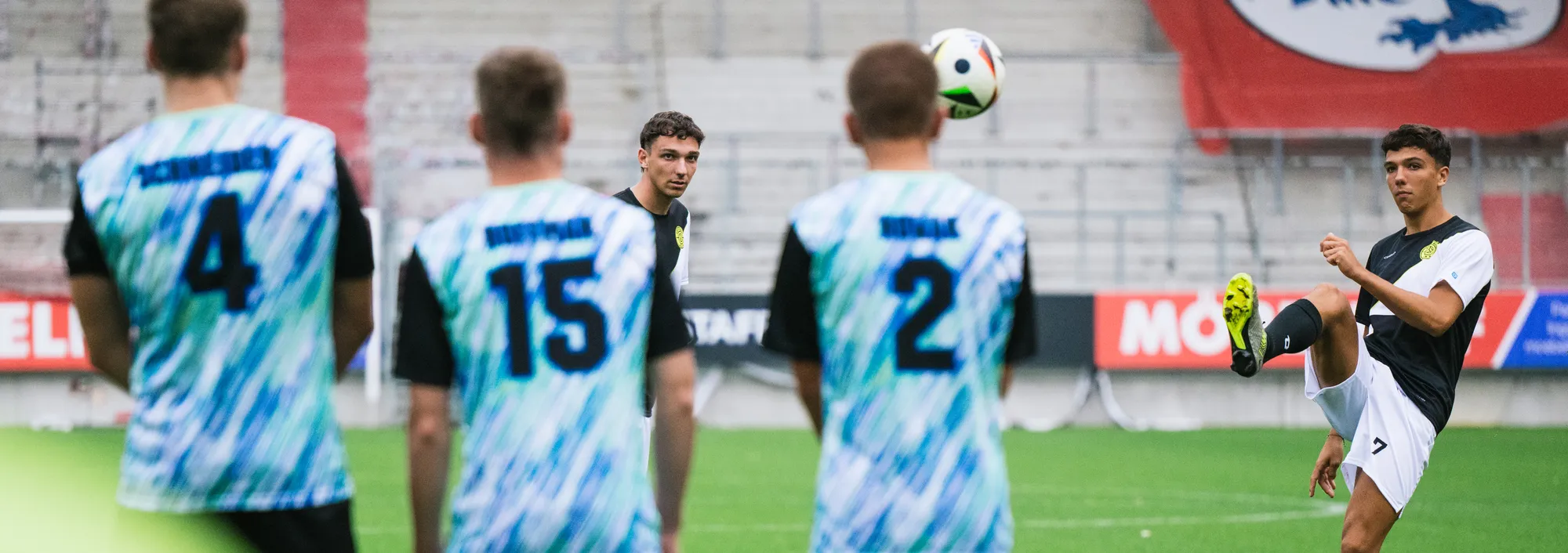 Eine Gruppe von Fußballspielern in bunten Trikots steht auf dem Spielfeld, während einer von ihnen einen Ball mit dem Fuß anhebt.