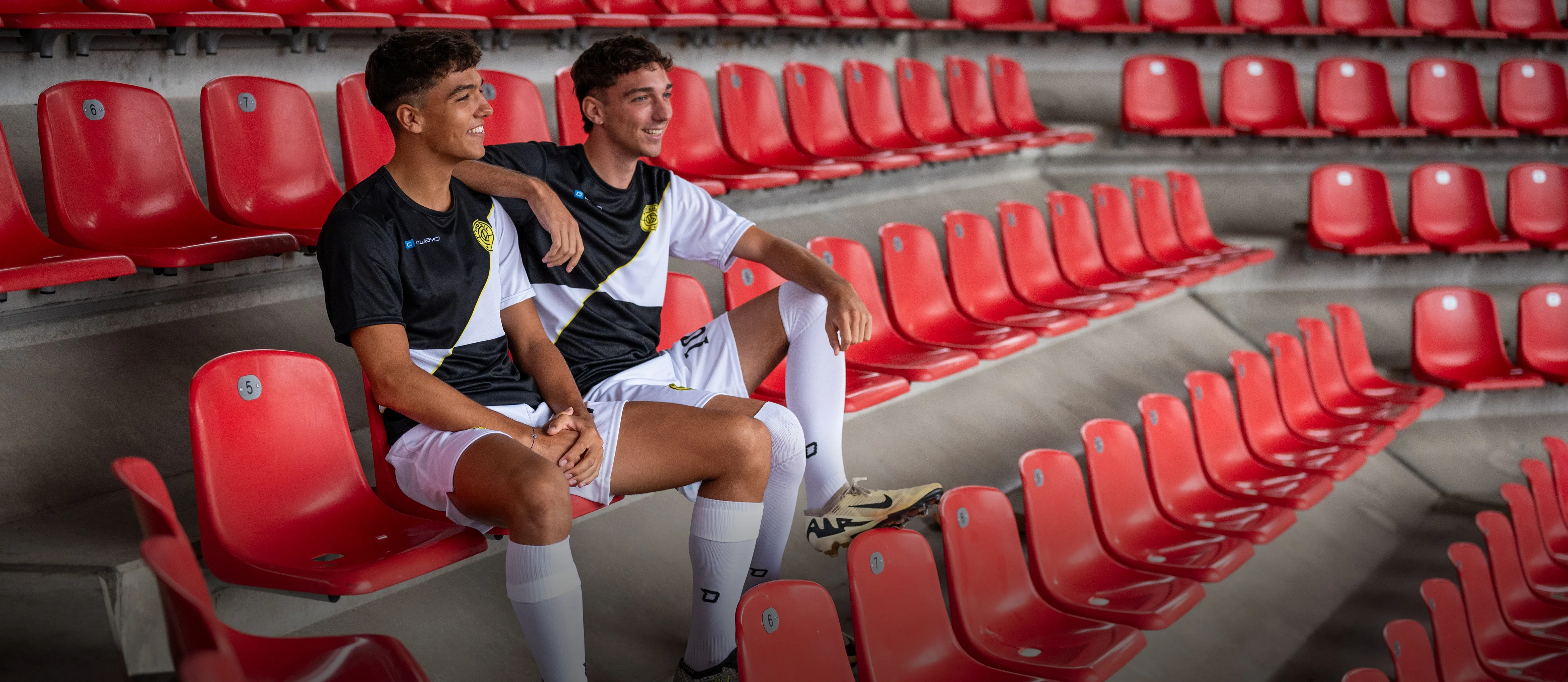 Zwei junge Männer in sportlicher Fußballkleidung sitzen lässig auf roten Stadionbänken.