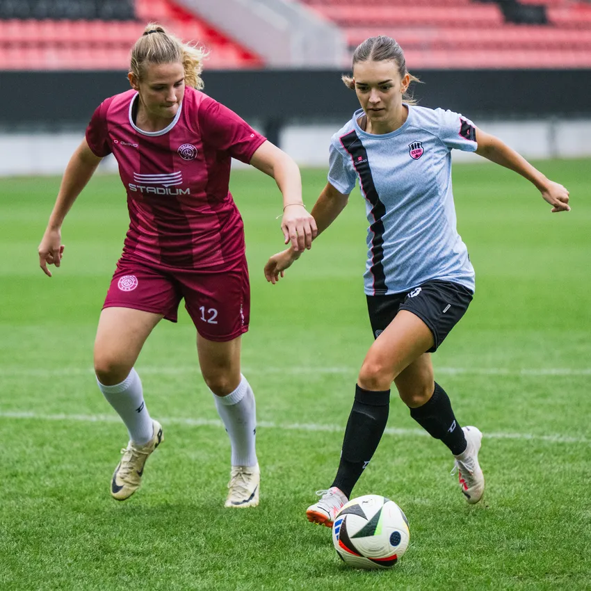 Zwei Frauen in Sportbekleidung spielen Fußball auf einem grünen Rasenplatz.
