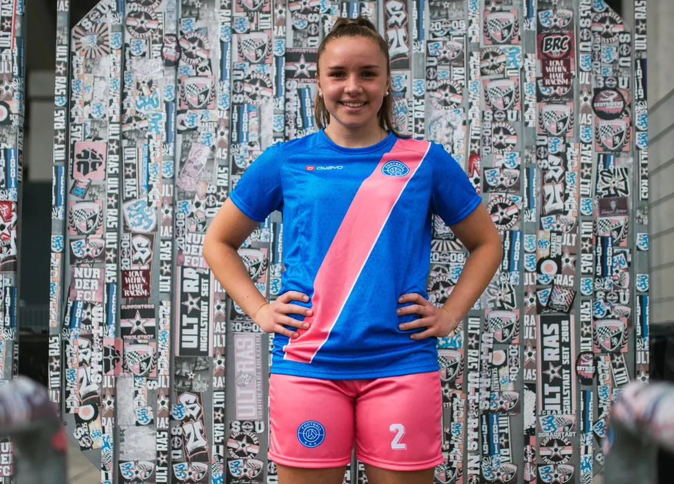 Eine junge Sportlerin posiert in einem blauen Fußballtrikot mit pinkem Streifen und pinken Shorts vor einer farbenfrohen Wand mit Aufklebern.