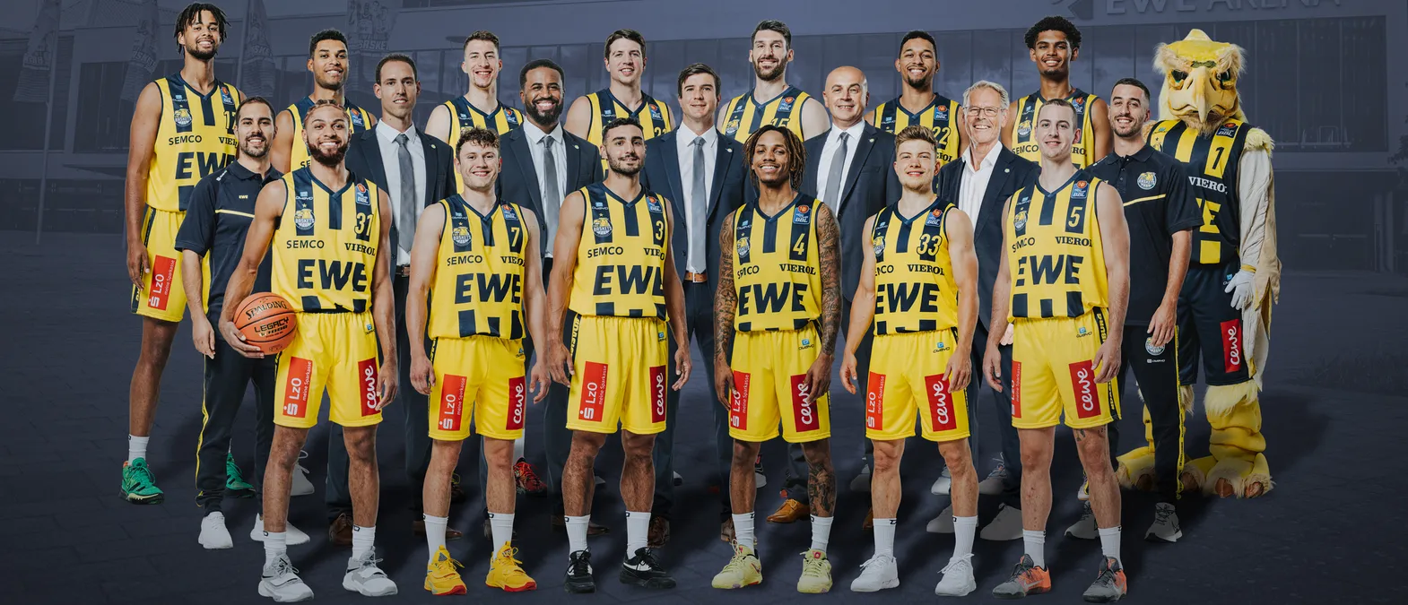 Eine Gruppenaufnahme eines Basketballteams in gelb-schwarzen Trikots mit den Sponsoren 'EWE' und 'SEMC', einem Maskottchen und einem Basketball, vor einem Gebäude.