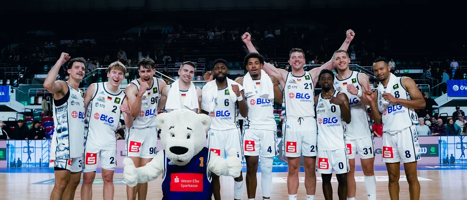 Basketballspieler in weißen Trikots mit dem Logo von BLG Logistics feiern auf dem Spielfeld, umgeben von einem Publikum, mit einem Eisbären-Maskottchen im Vordergrund.