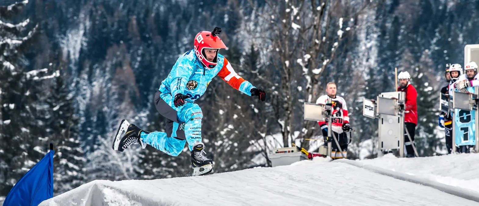 Ein Eishockeyspieler in einer blau-weißen, mit Sponsorenlogos bedruckten Trikot und einer roten Helm springt über eine Schneewelle auf einer Eisbahn, während im Hintergrund weitere Spieler und Bäume zu sehen sind.