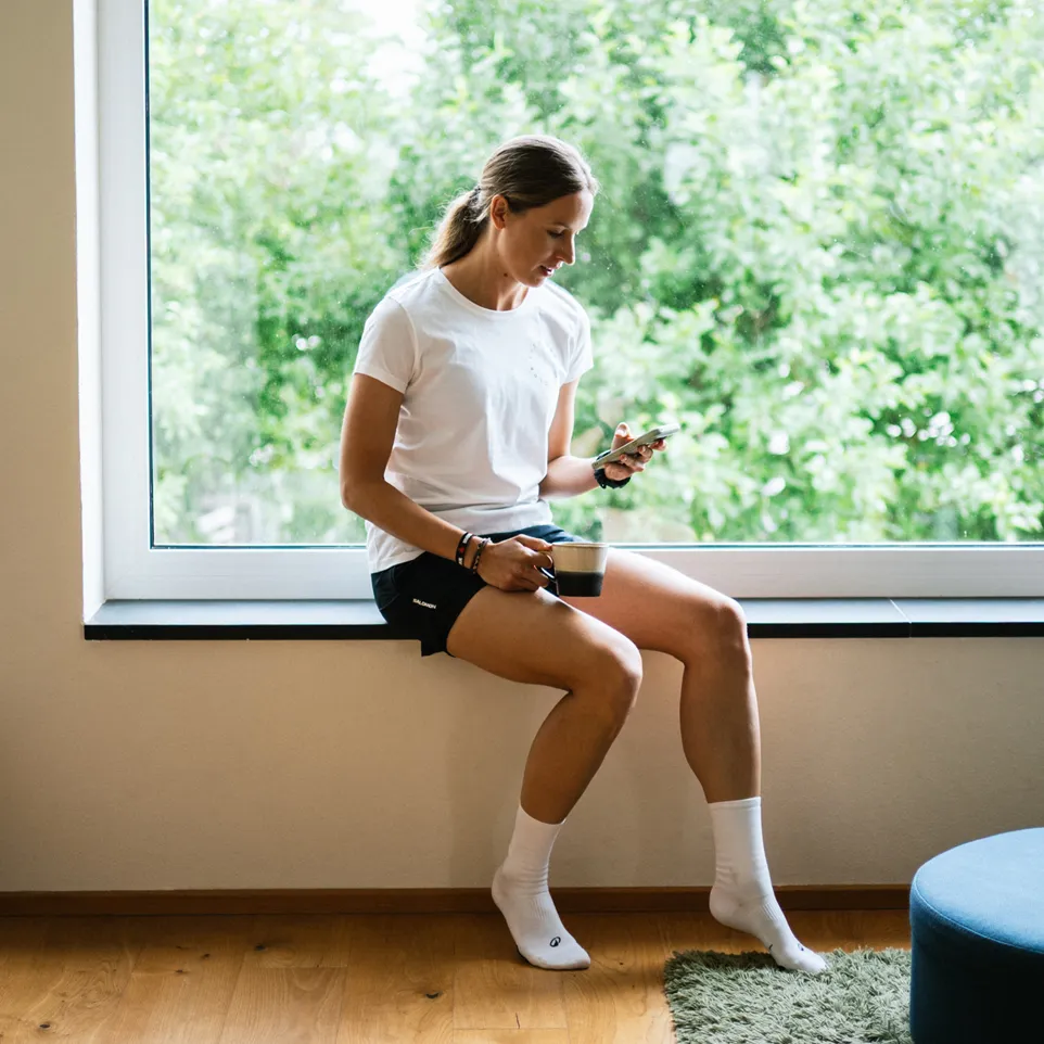 Eine Frau sitzt auf einer Fensterbank, trägt ein weißes T-Shirt und schwarze Shorts, während sie ein Smartphone in der Hand hält und eine Tasse Kaffee genießt.