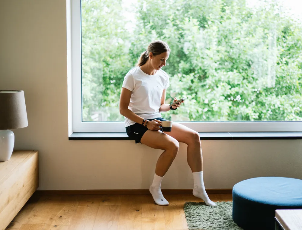 Eine Frau sitzt auf einem Fensterbrett und schaut auf ihr Handy, während sie eine Tasse Kaffee hält, gekleidet in ein weißes T-Shirt und schwarze Shorts.