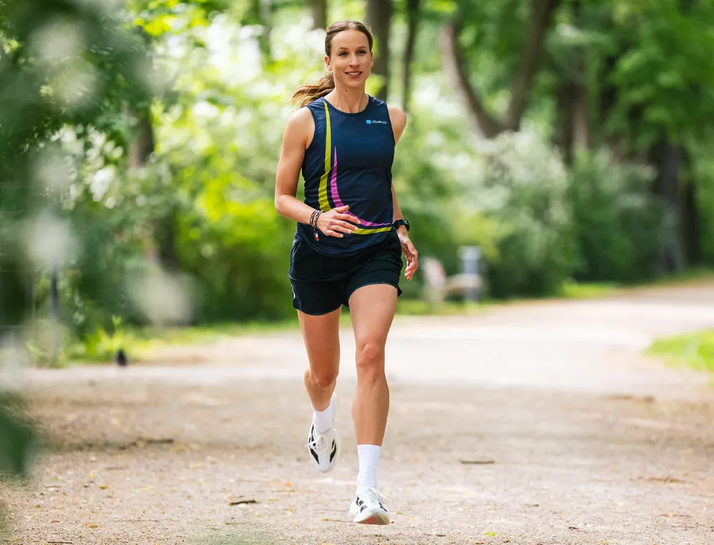 Eine sportliche Frau läuft auf einem schmalen Weg durch einen grünen Park, gekleidet in einem blauen Trikot mit bunten Akzenten und einer kurzen Hose.