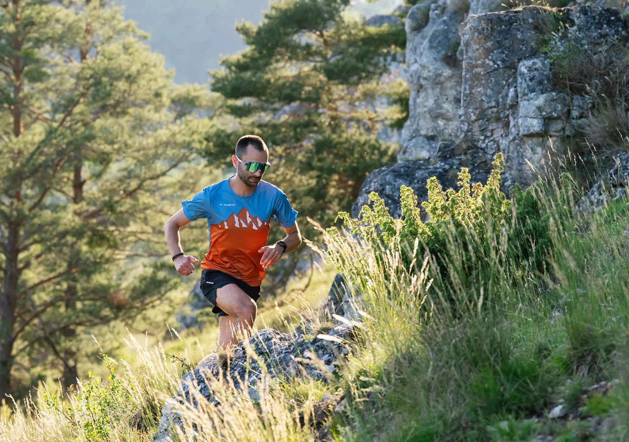 Ein Läufer in einer bunten Sportbekleidung, der über felsige Terrain in einem bewaldeten Gebiet läuft.