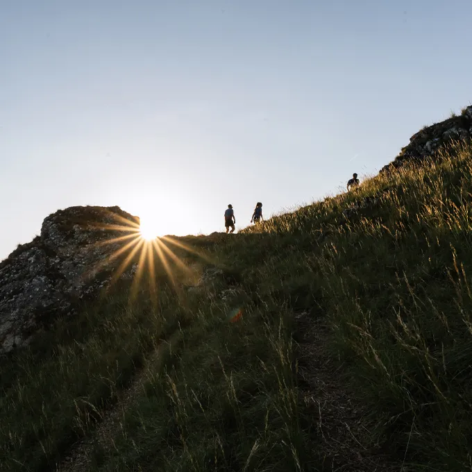 Zwei Wanderer steigen einen bewachsenen Hügel bei Sonnenuntergang hinauf, während die Sonne hinter einem Felsen strahlt.