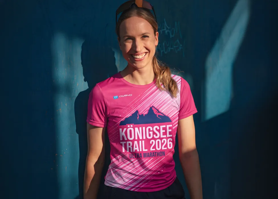 Eine lächelnde Frau trägt ein pinkes T-Shirt mit dem Aufdruck 'Königsee Trail 2026 Ultra Marathon' vor einer blauen Wand.