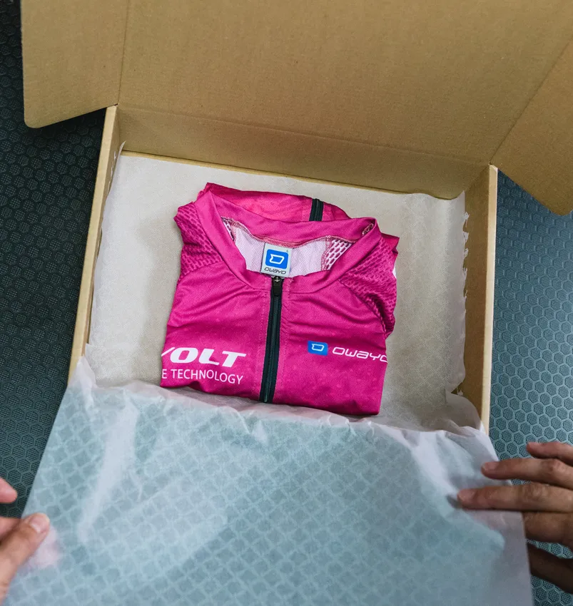 Eine gefaltete, pinkfarbene, maßgeschneiderte Sportbekleidung von owayo liegt in einer geöffneten Pappschachtel auf weißem Papier, die von Händen gehalten wird.