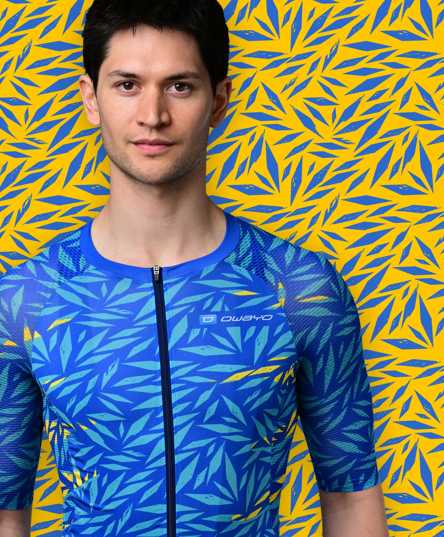 Ein Mann trägt ein blaues, kurzärmeliges Sporttrikot mit einem blauen und gelben Blattmuster, das an eine tropische Pflanze erinnert, vor einem gelben Hintergrund mit blauen Blattmustern.