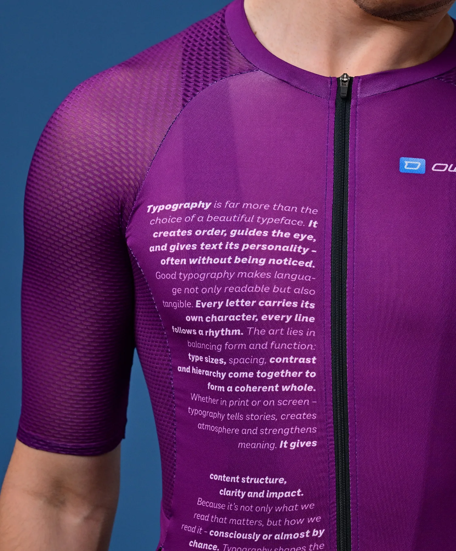 Ein Mann trägt ein lila, kurzärmeliges Sporttrikot mit einem Text über Typografie auf der Brust und einem kleinen Logo auf der Schulter.