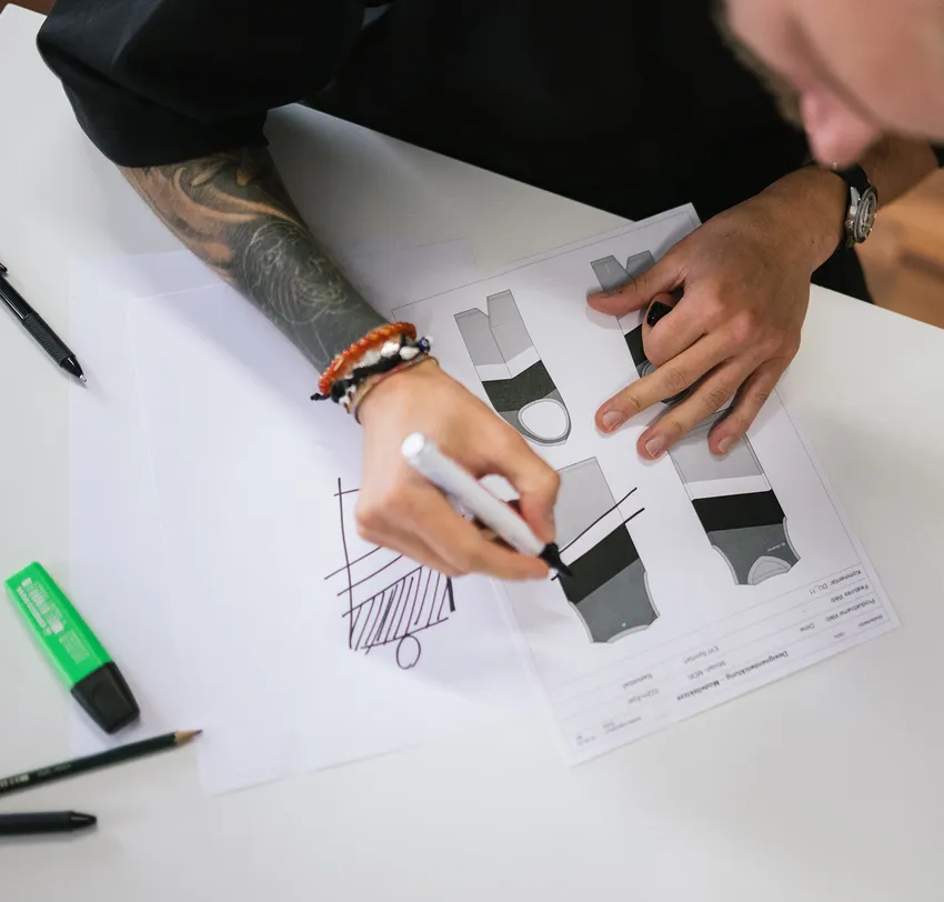 Eine Person mit tätowiertem Arm entwirft mit einem Marker auf einem Blatt Papier Designs für maßgeschneiderte Sportbekleidung.