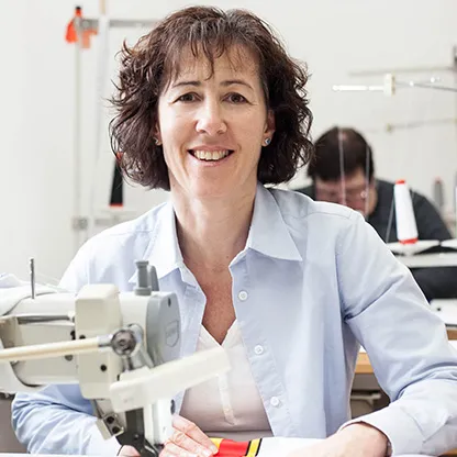 Eine Frau lächelt in die Kamera, während sie an einer Nähmaschine in einer Werkstatt arbeitet, wobei im Hintergrund eine weitere Person an einer Nähmaschine zu sehen ist.