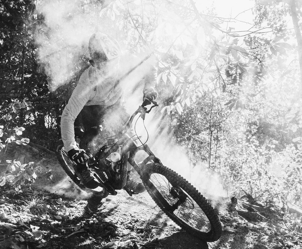 Ein Mountainbiker in einer dynamischen Bewegung auf einem Waldweg, umgeben von Staub und Lichtstrahlen, trägt eine lange Ärmel-Oberteil von owayo.